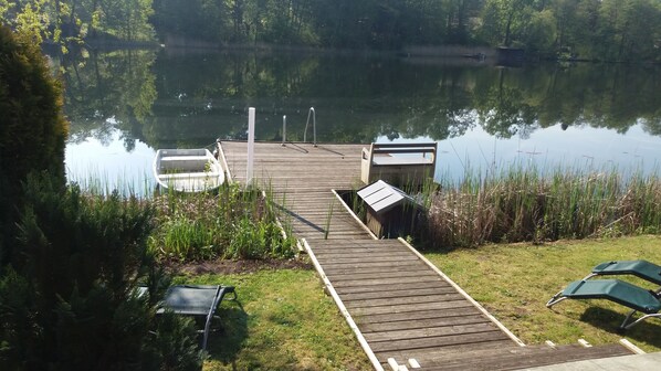 A wooden dock extending into a calm lake, surrounded by lush greenery and a grassy area with a bench and a small boat. - Haus Kathrin (Wendisch Rietz)