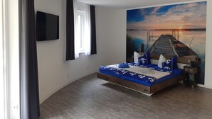 Een slaapkamer met een bed, een schilderij van een zonsondergang, een plafondlamp en een raam met gordijnen.