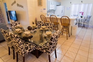 Un comedor con una mesa de vidrio rodeada de sillas estampadas, una cocina al fondo y un cubo de basura al lado.