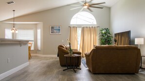A spacious living area with a high ceiling fan, a large window with curtains, a brown sofa, a small round table with a plant, and a painting on the wall. - Casablanca Villaview by Orlando Short Term (Kissimmee)