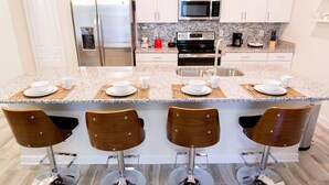 A modern kitchen with a granite countertop, stainless steel appliances, and four wooden bar stools. - Gatsby Storey by Orlando Short Term (Kissimmee)