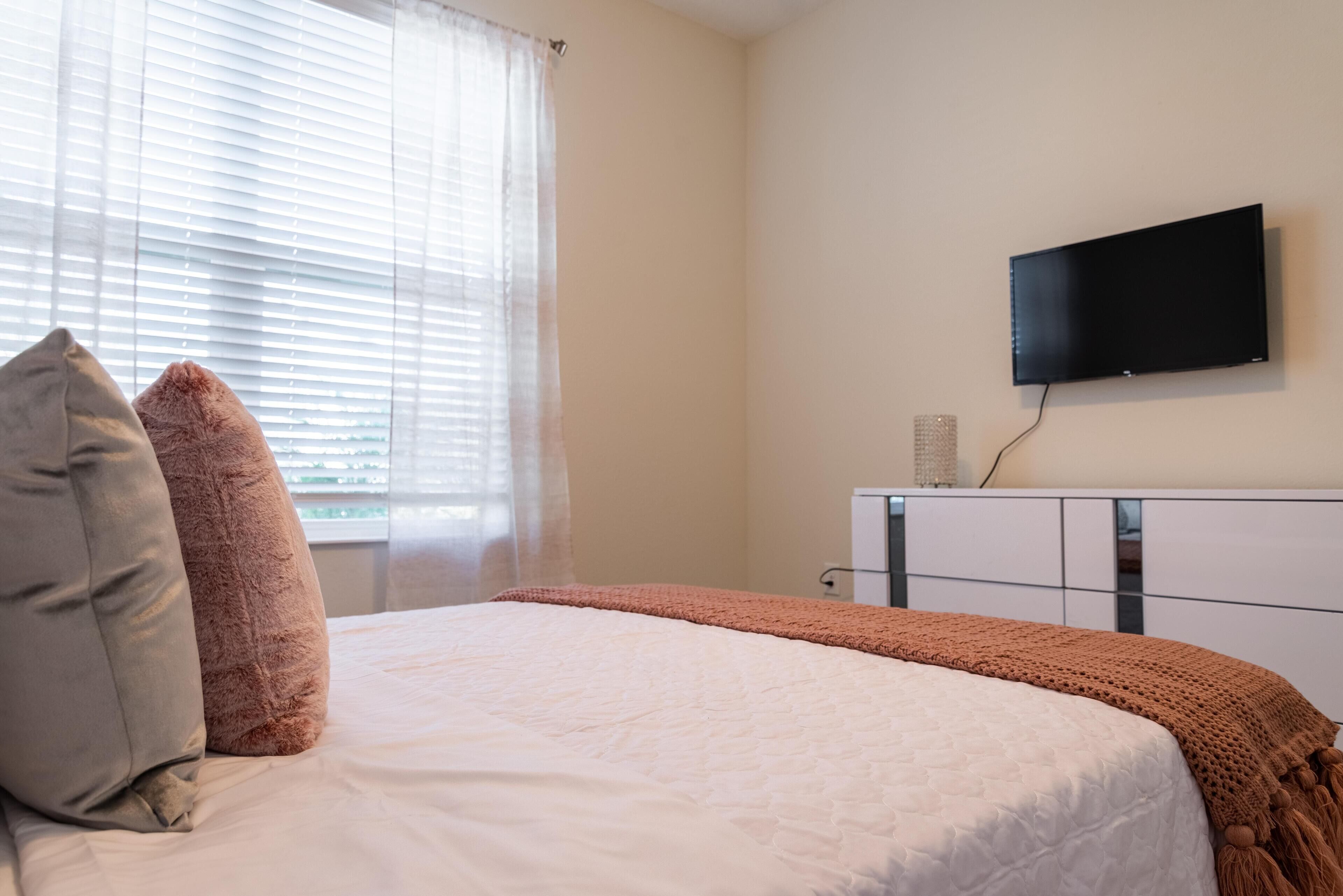 A bedroom with a bed, pillows, a TV mounted on the wall, and a window with blinds.