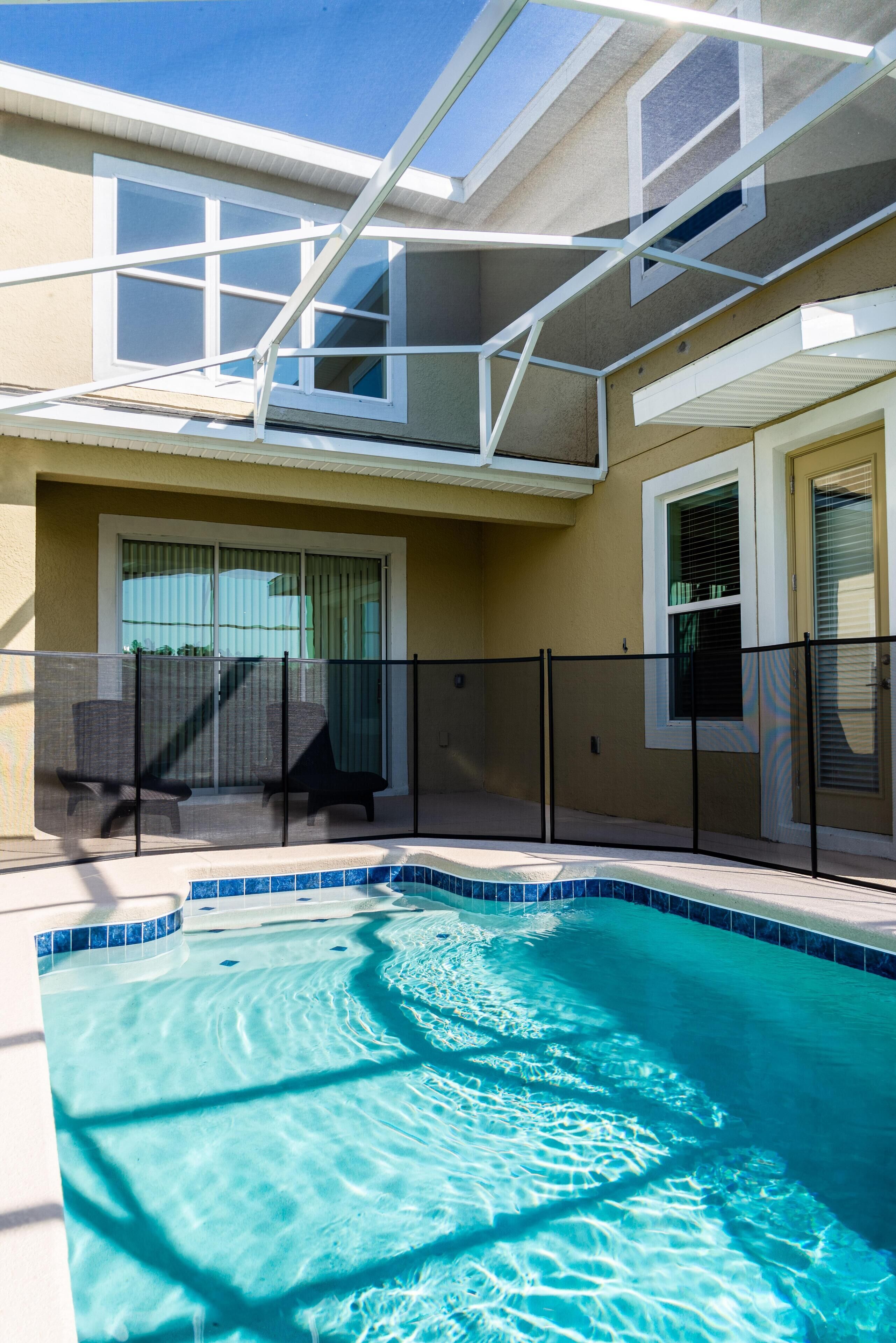 A swimming pool with a clear blue water surface, a covered patio area with chairs, and a building with a beige exterior and white trim.