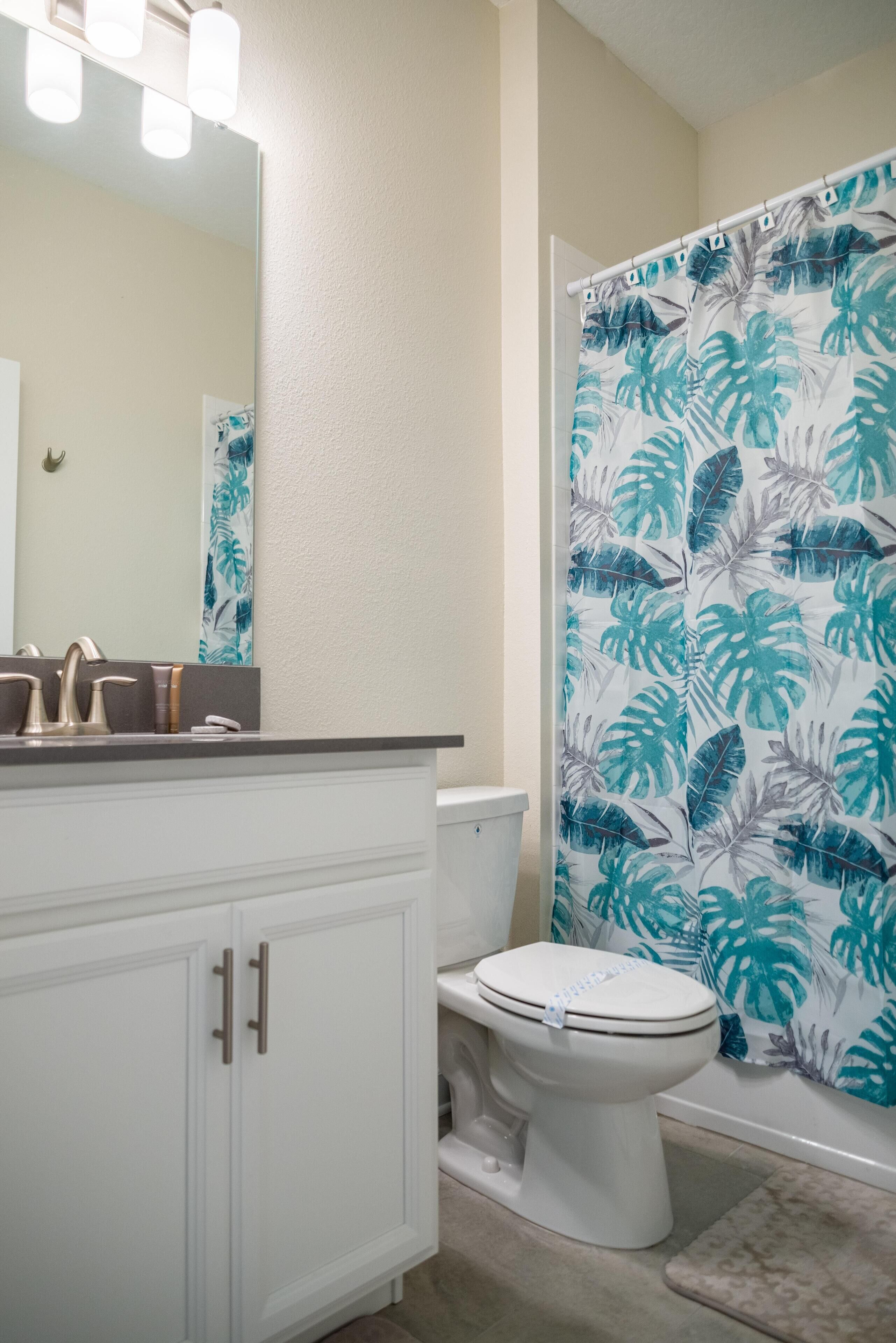 A bathroom with a white toilet, a vanity with a mirror, and a shower curtain with a tropical leaf pattern.