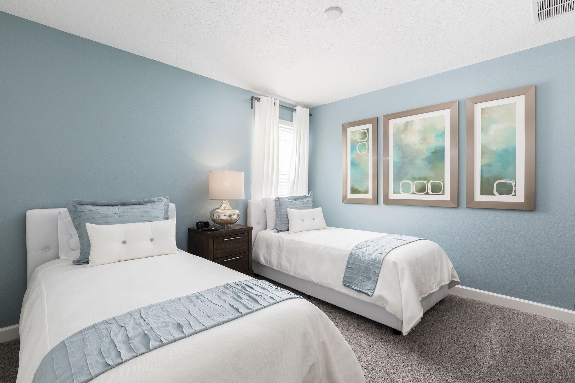 A bedroom with two beds, a nightstand, a lamp, and three framed artworks on the wall.