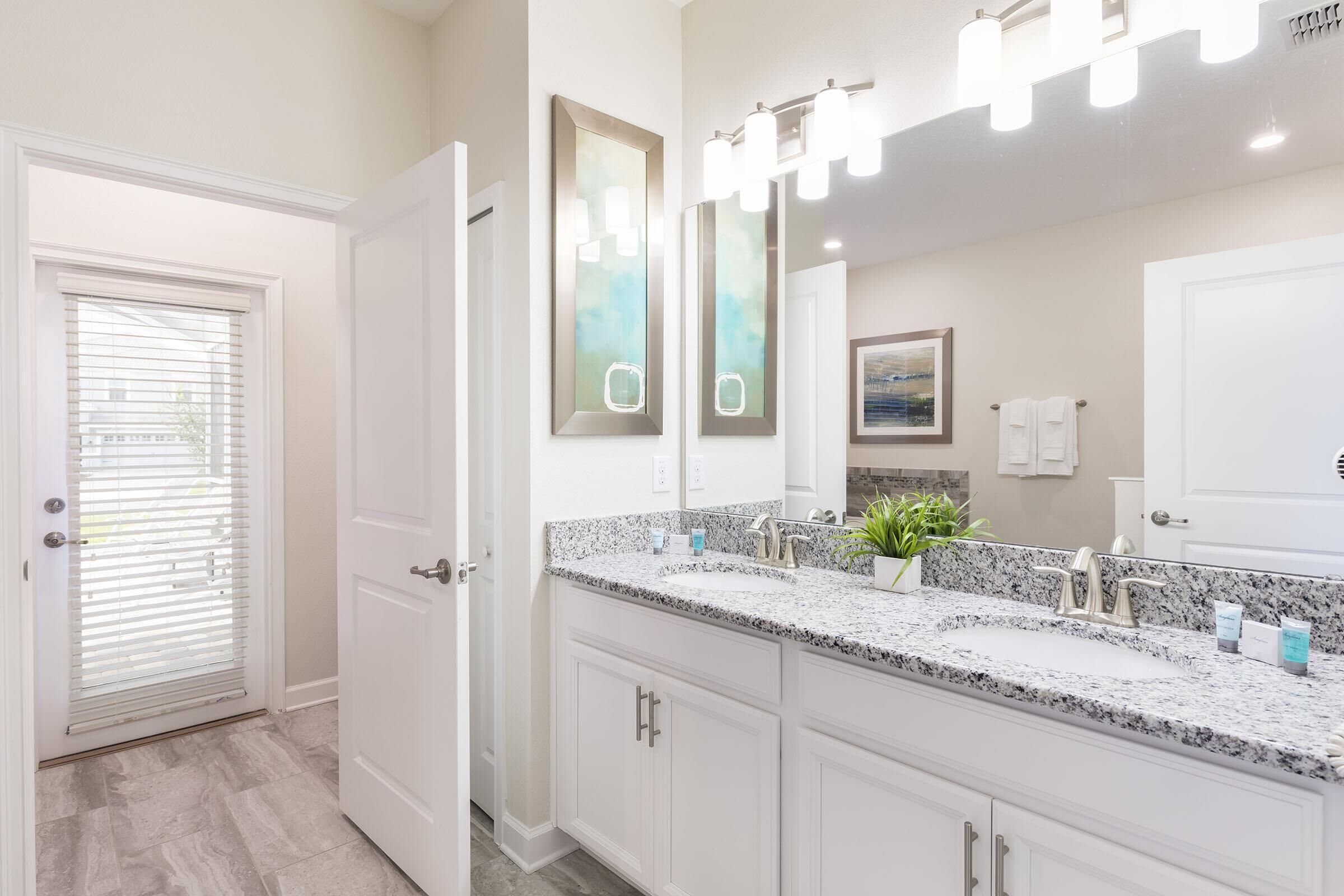 A modern bathroom with a double sink vanity, a large mirror with lights, and a door leading to another room.