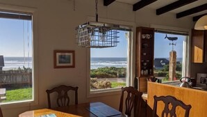A dining area with a wooden table and chairs, a hanging light fixture, and a view of the ocean through large windows. - Pacific Pearl at Heceta Beach  - dog friendly (Florence)