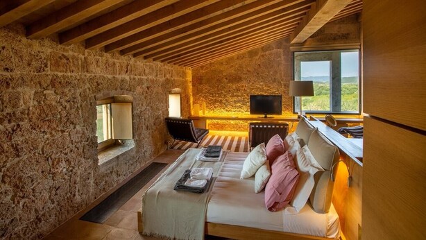 Ein rustikaler Raum mit Steinwänden und Holzbalkendecke, ein Bett mit Kissen, ein Fernseher und ein Fenster mit Blick.