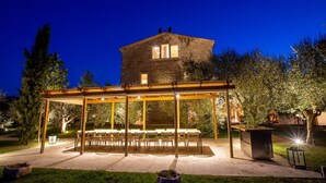 Ein Terrassenbereich im Freien mit einer Pergola, Tischen und Stühlen, umgeben von Bäumen und einem Steingebäude im Hintergrund.