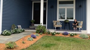 A house with a blue exterior, a covered porch with two chairs, and a small garden with various plants and flowers. - Industrial chic farmhouse..... (Oshkosh)