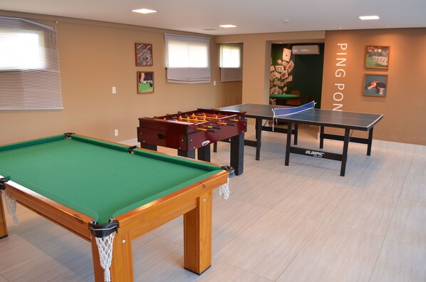 A pool table, foosball table, ping pong table, and a foosball table in a room with beige walls and windows.