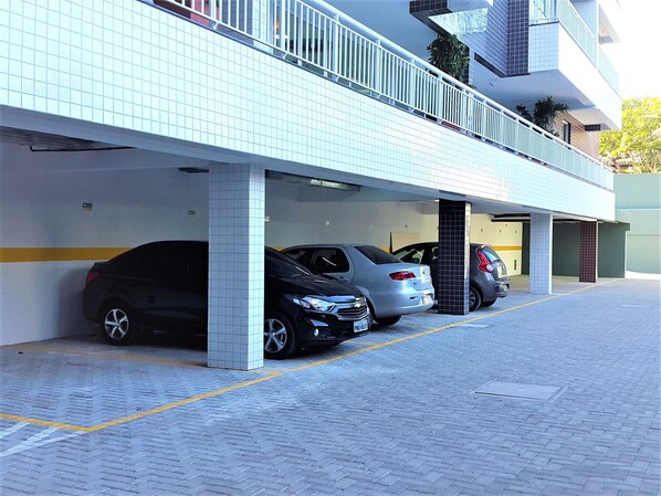 A parking garage with multiple cars parked under a building with tiled walls and a balcony. - Beachfront Condominium BEACH WAY - PORTO DAS DUNAS - CE, APARTMENT WITH SEA VIEW (Aquiraz)