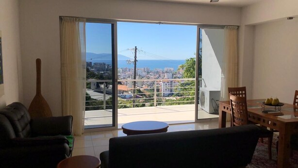 Una sala de estar con un sofá, una mesita redonda y una mesa de comedor con sillas. Hay un ventanal con cortinas que ofrece vistas a un paisaje urbano y montañas.