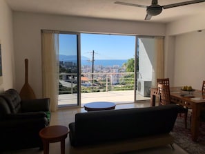 Un salon comprenant un canapé, une petite table ronde et une table à manger avec des chaises. Une grande fenêtre, ornée de rideaux, offre une vue sur le paysage urbain et les montagnes.