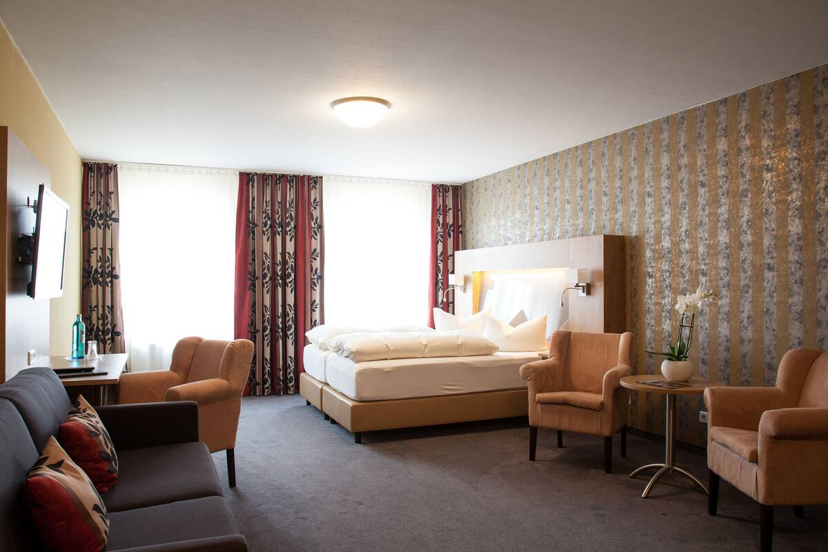 A hotel room with a bed, two armchairs, a sofa, a small table with a vase, and a television on the wall.