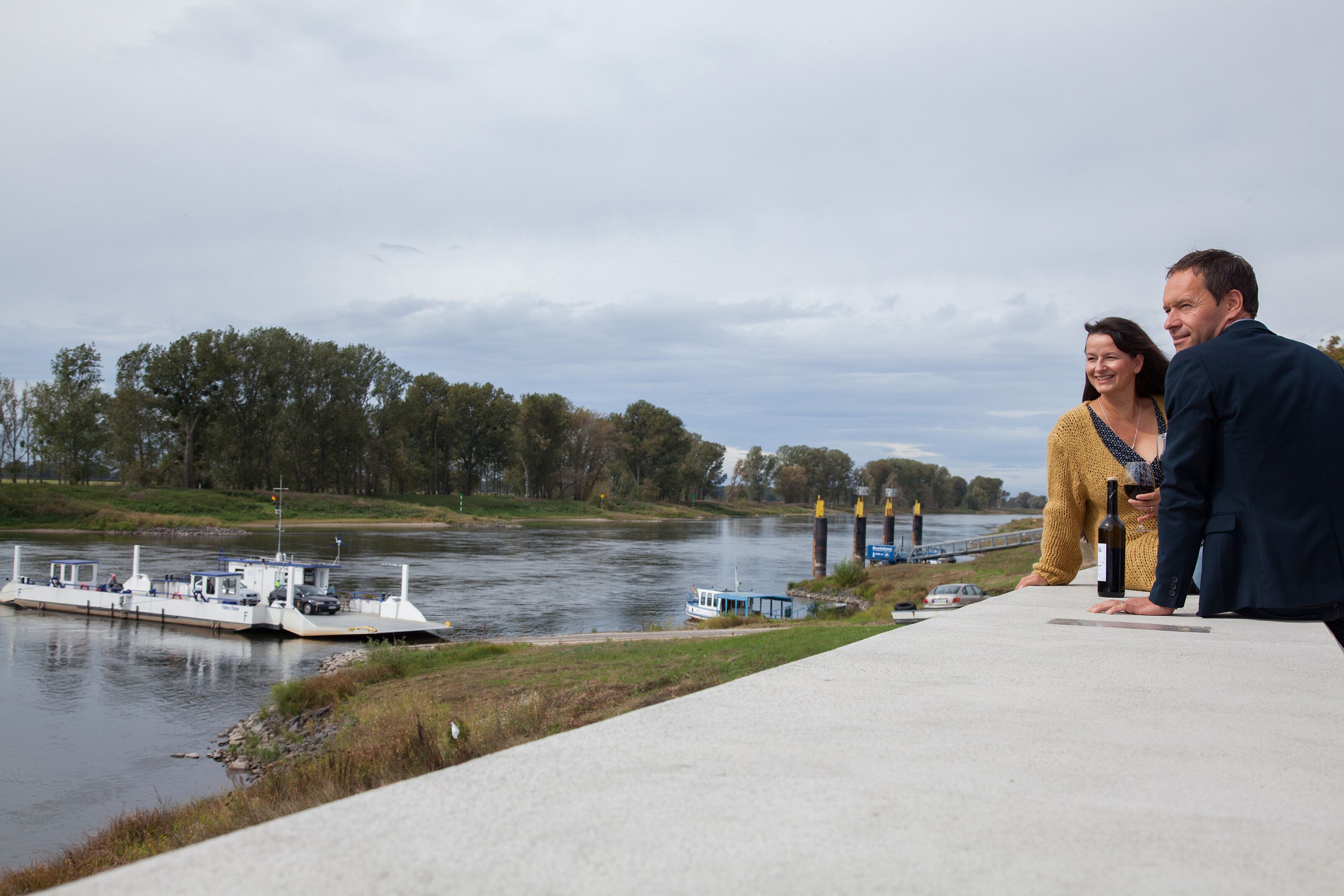 A river with a boat docked on the left, a bridge in the background, and a concrete walkway in the foreground.