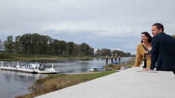 A river with a boat docked on the left, a bridge in the background, and a concrete walkway in the foreground.