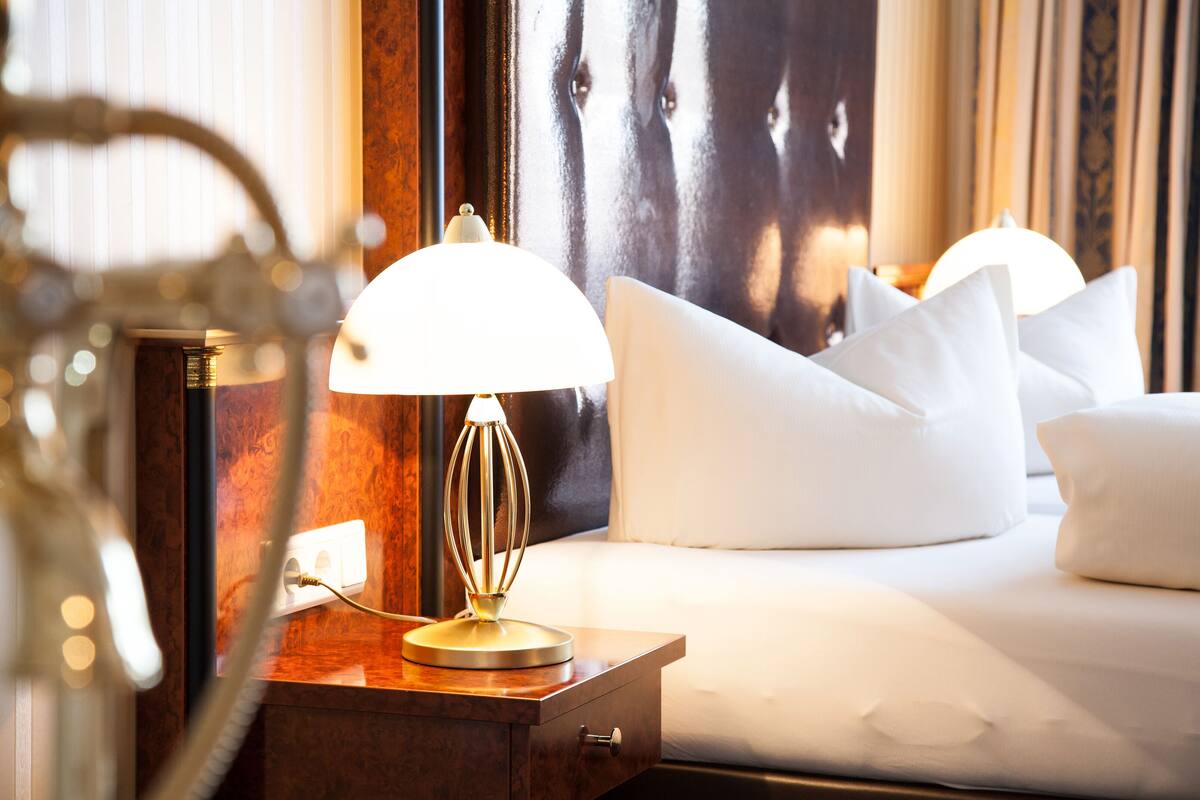 A neatly made bed with white linens and pillows, a bedside lamp, and a wooden nightstand with a power outlet.