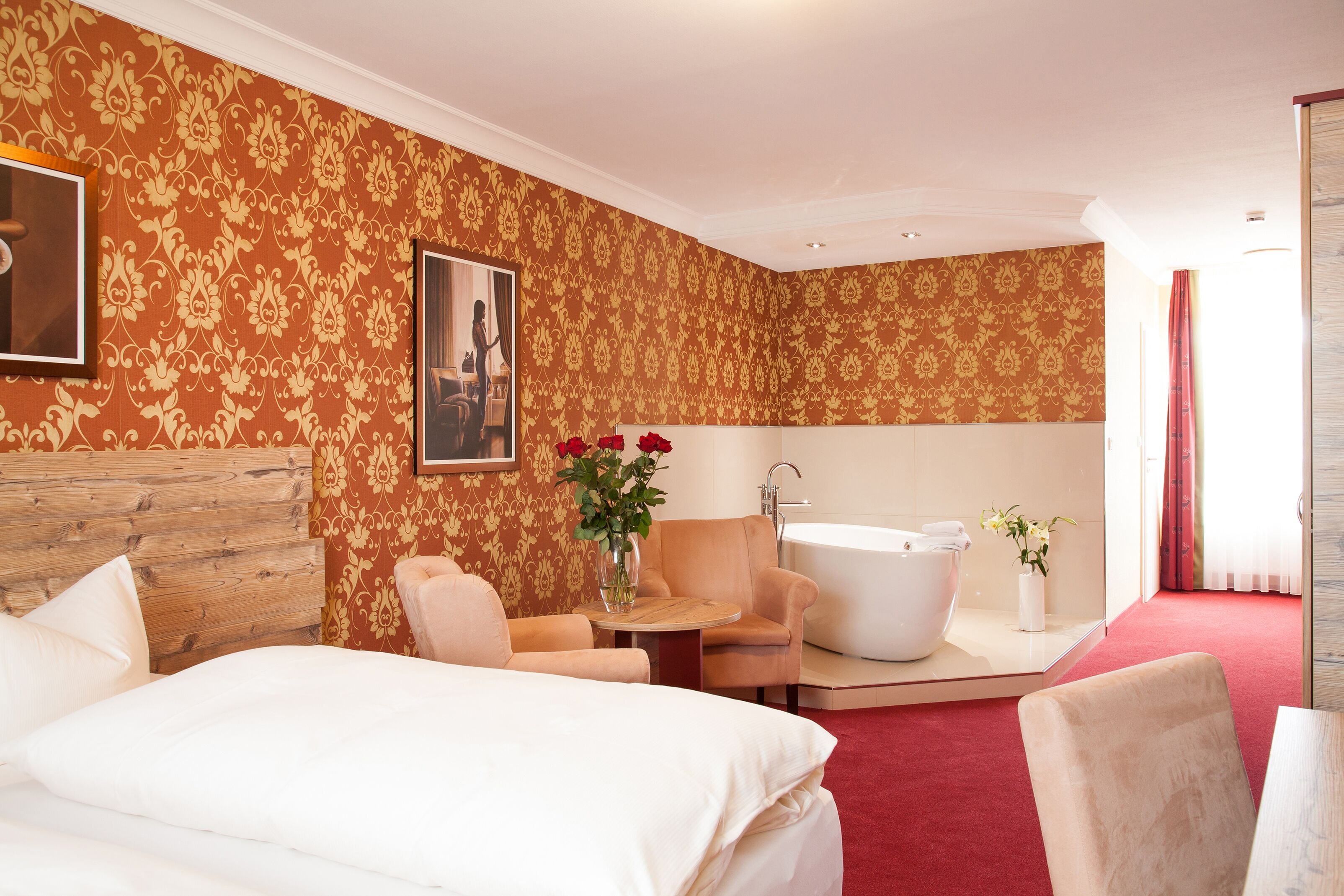 Habitación de hotel con cabecera de madera, cama, sofá, una mesita, un florero con flores, bañera y espejo.