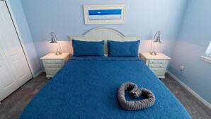 En snyggt bäddad säng med ett blått överkast och kuddar, två sänglampor och en inramad tavla på väggen.