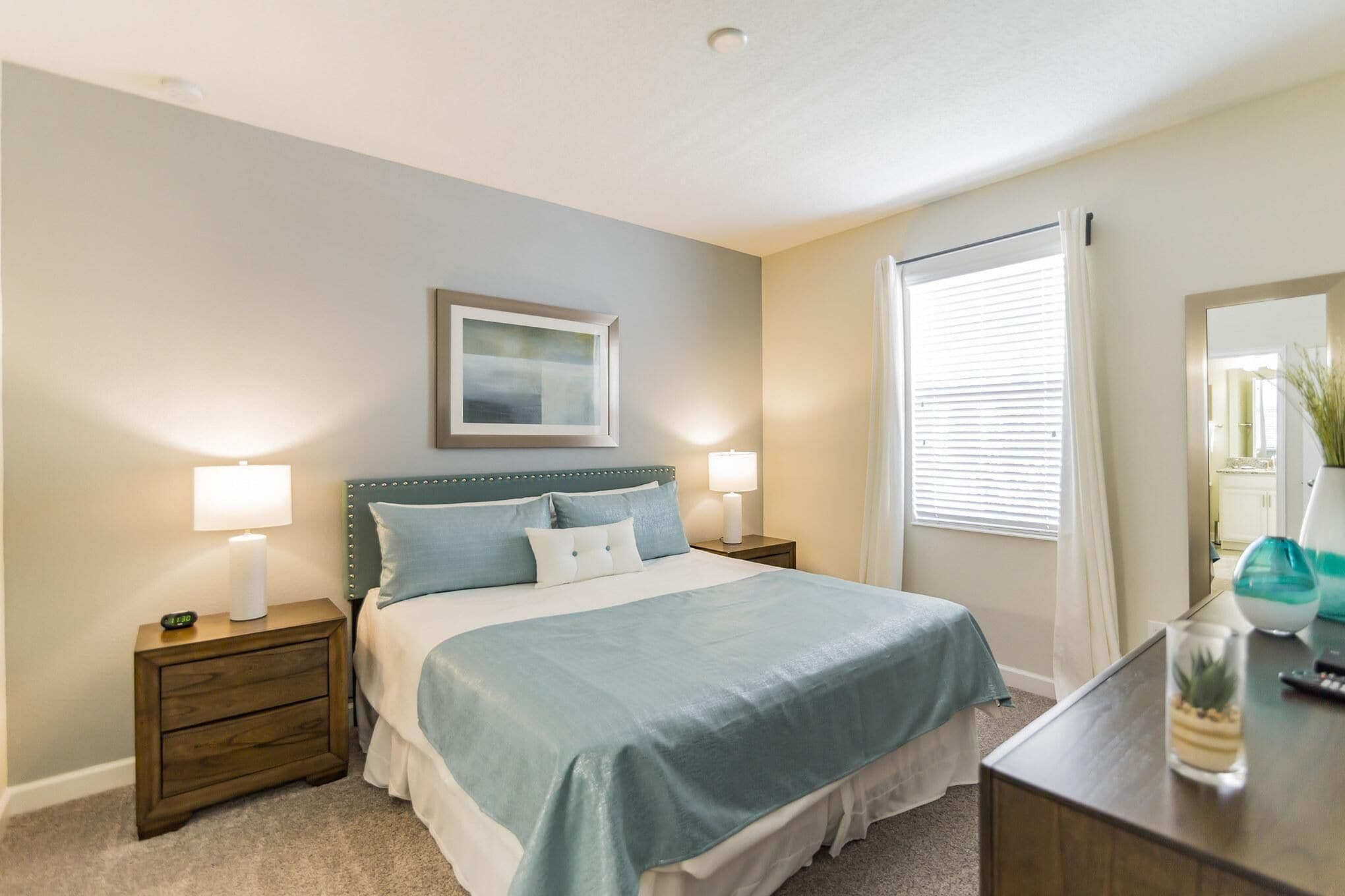 A bedroom with a bed, two bedside tables with lamps, a nightstand, a dresser, a mirror, and a window with blinds.