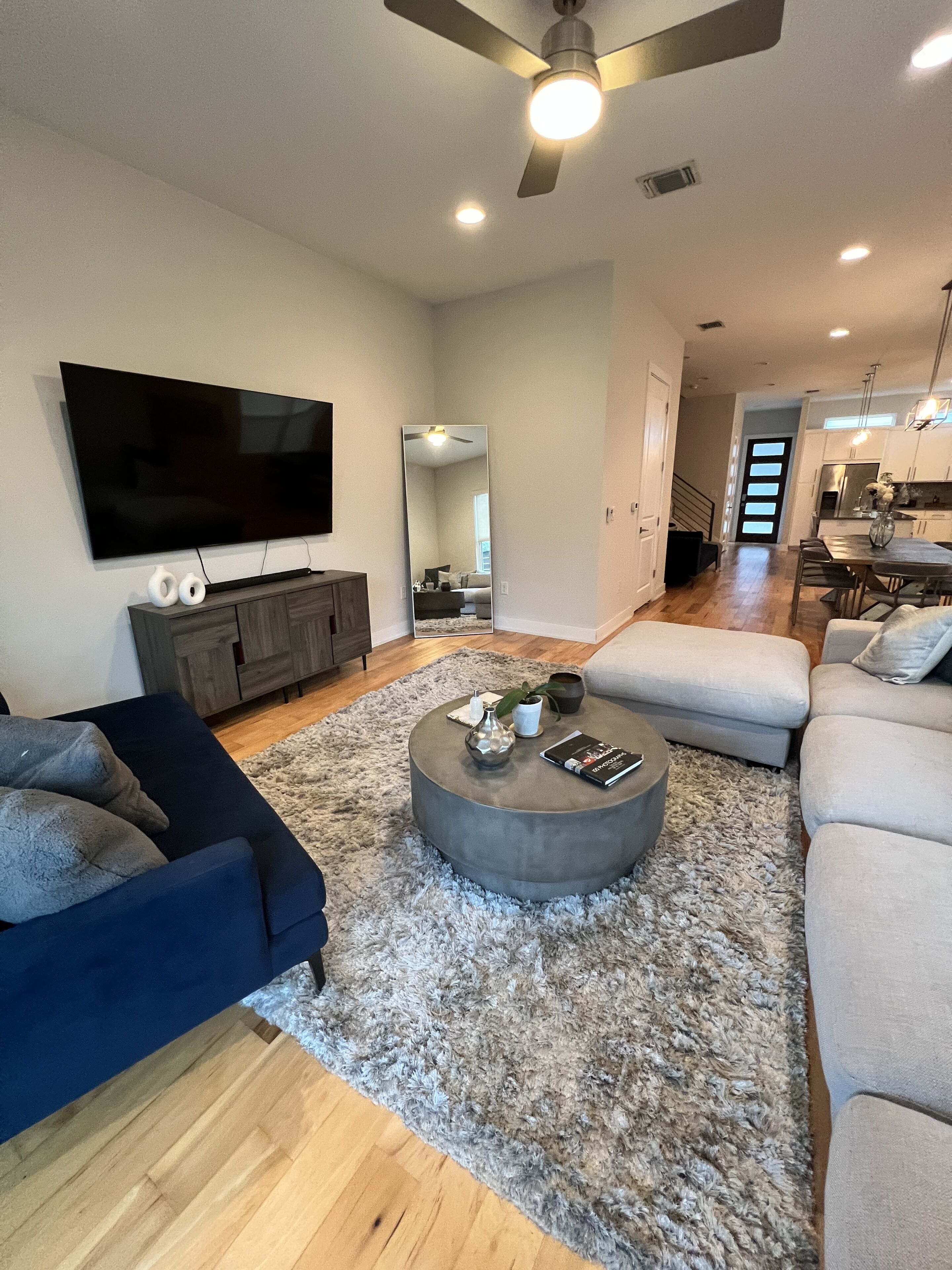 A modern living room with a flat-screen TV, dark blue sofa, round coffee table, and a large mirror.