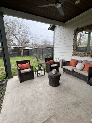Relax in this serene outdoor space with wicker furniture and a ceiling fan overhead, surrounded by a grassy yard and a building in the background.