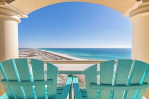 Un balcón con sillas Adirondack turquesas que da a una playa bajo un cielo azul claro.