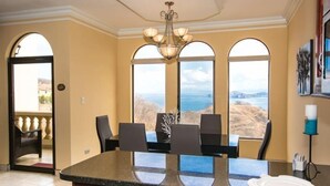 A dining area with a large island, chairs, and a view of the ocean through arched windows. - Casa Q (Sardinal)