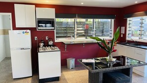A compact kitchen with a refrigerator, stove, and sink, a window with blinds, and a small table with a plant.