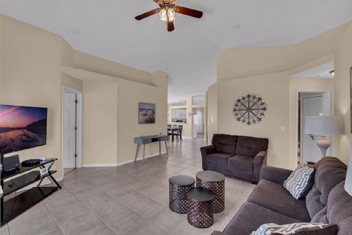 A spacious living room with a large flat-screen TV, a brown sofa, two patterned side tables, and a ceiling fan.
