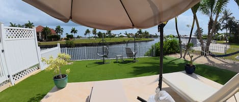 A patio with a white lounge chair, a beige umbrella, and a view of a water body and greenery.
