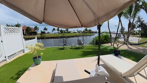A patio with a white lounge chair, a beige umbrella, and a view of a water body and greenery.
