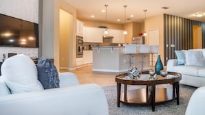 A modern living room with a white sofa, a coffee table with decorative items, and a kitchen area with white cabinets and a central island. - Sunrise Valley Villa by Orlando Short Term (Kissimmee)