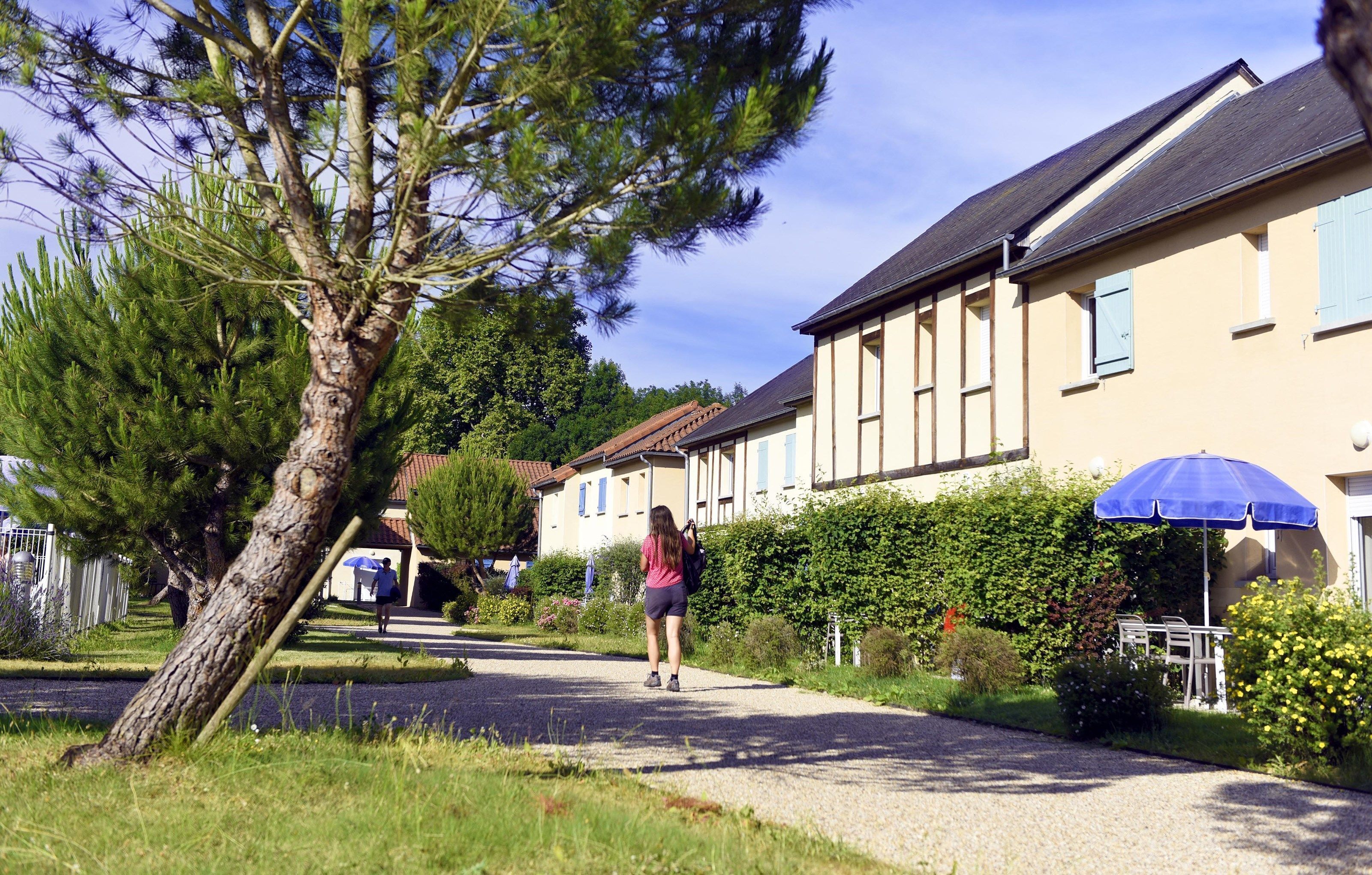 Une rue résidentielle bordée de maisons, où l’on voit une personne qui marche et un parapluie bleu.