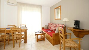 A cozy living room with a wooden sofa, a dining table with chairs, a coffee machine, and a large window with curtains. - Maisonnette Abordable 5mn de Lascaux (Montignac)