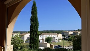 A view through an arched doorway of a residential area with houses, a tall tree, and a clear blue sky. - Appart Abordable avec Accès à 2 Piscines! (Salavas)