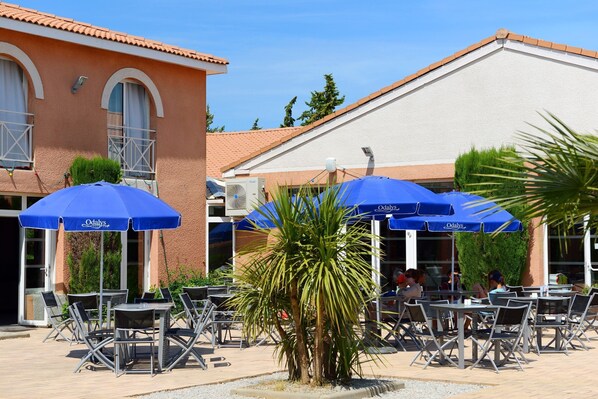 An outdoor seating area with tables and chairs under blue umbrellas, a palm tree, and a building with a tiled roof.