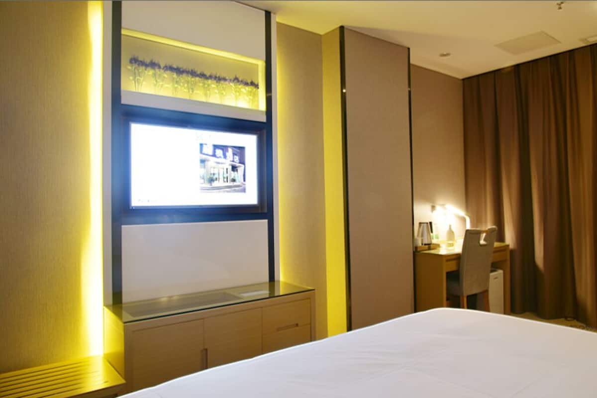 Quarto de hotel com cama, escrivaninha com cadeira, mesinha, luminária e janela com vista para edifícios.
