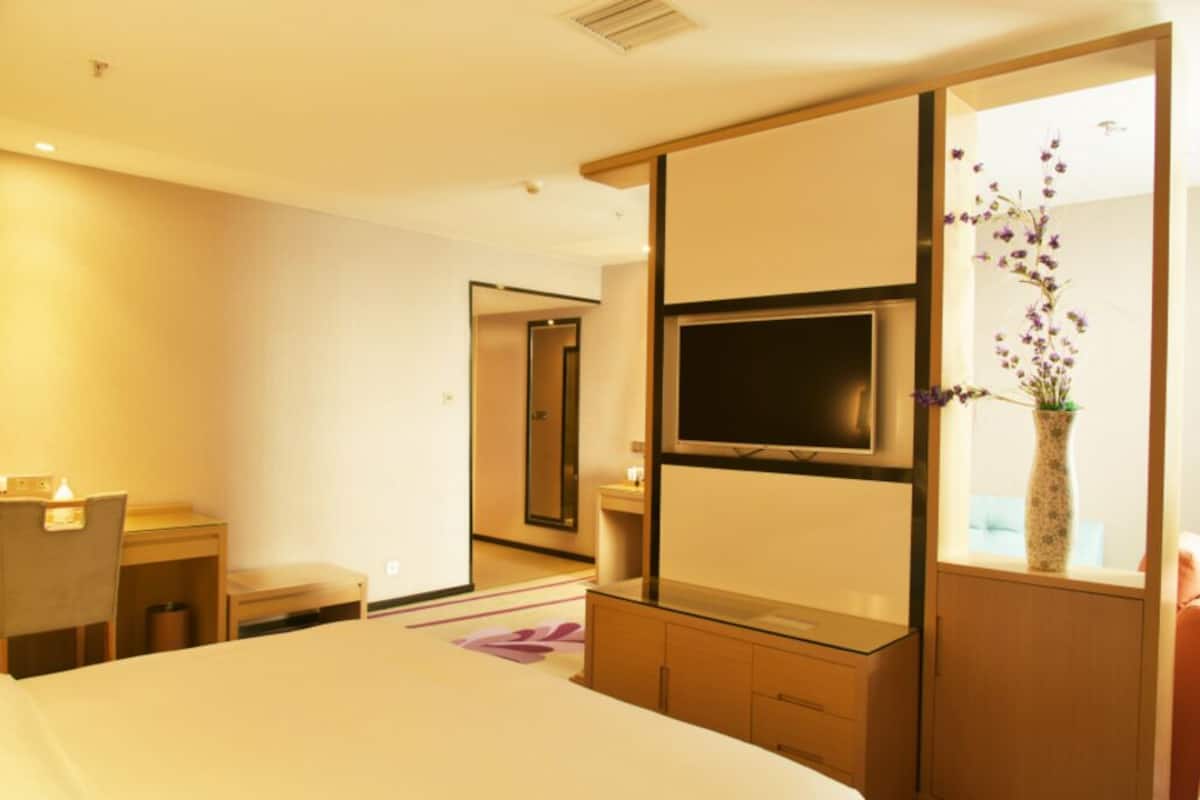 Quarto de hotel com cama, escrivaninha, televisão, espelho e um vaso com flores.