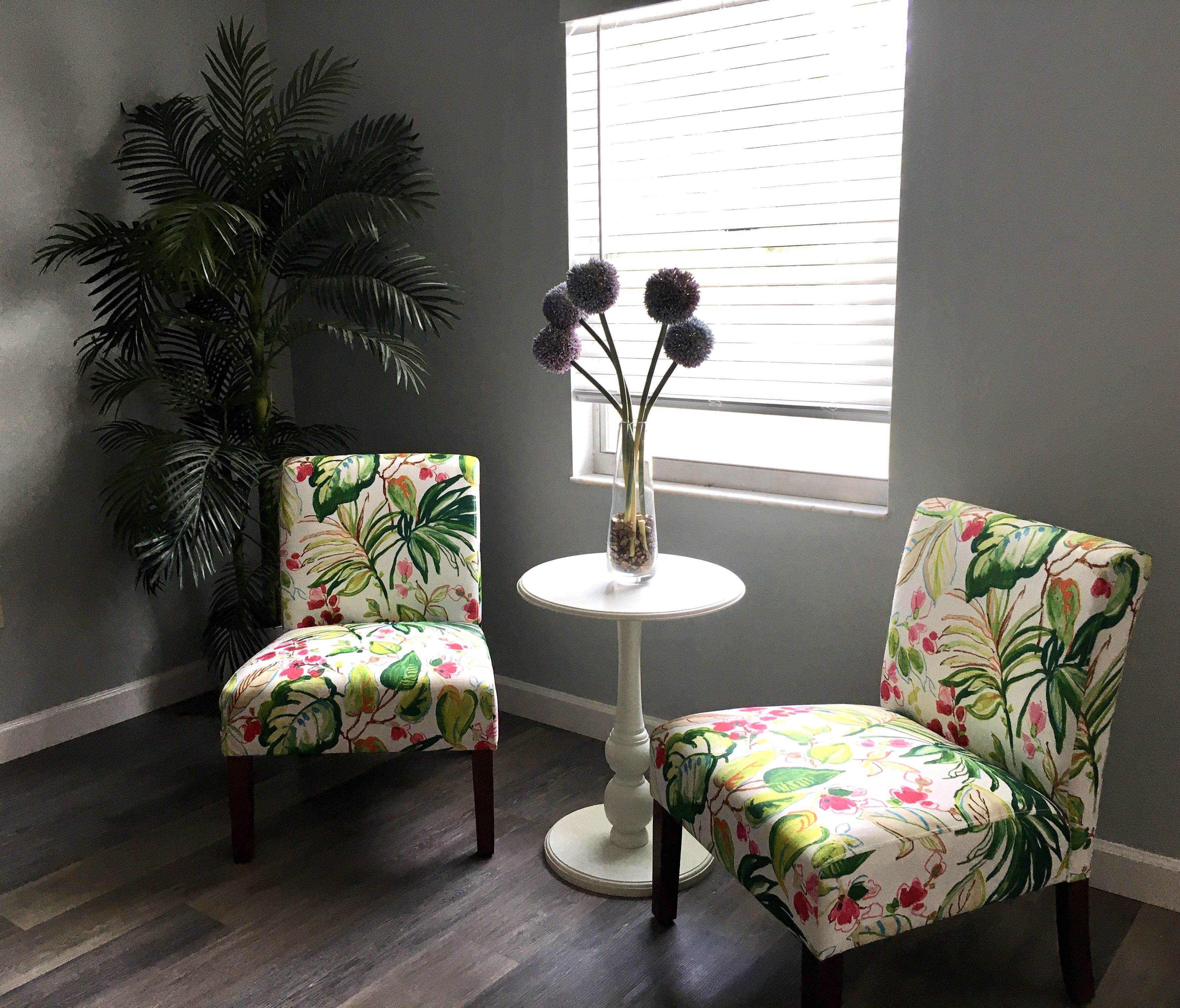 Ein Zimmer mit zwei floralen Sesseln, einem kleinen runden Tisch mit einer Vase und einer großen Küstenpflanze.