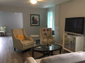 Un salon avec deux fauteuils, une table basse en verre, un téléviseur posé sur un meuble et un tableau accroché au mur.
