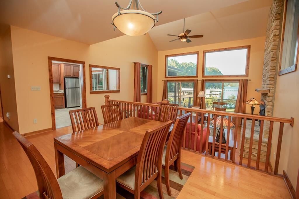 Un comedor con una mesa y sillas de madera, un ventilador de techo y ventanas amplias con vistas al exterior.