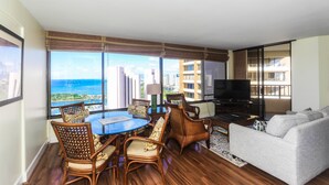 Ein geräumiges Wohnzimmer mit großem Fenster und Blick auf Stadt und Meer, einem runden Esstisch mit Stühlen, einer Couch und einem Flachbildfernseher.