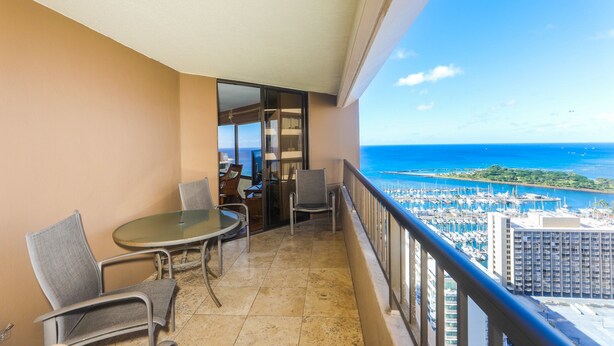 Ein Balkon mit Glastisch und Stühlen, Blick auf das Meer und den Yachthafen, sowie ein Gebäude im Hintergrund.