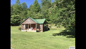 Une petite cabane en bois, surmontée d’un toit vert, entourée d’un champ herbeux et d’arbres.