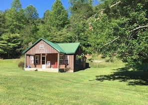 Une petite cabane en bois, surmontée d’un toit vert, entourée d’un champ herbeux et d’arbres.