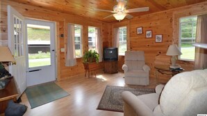 L’intérieur d’un chalet accueillant, avec des murs en bois, un ventilateur de plafond, un téléviseur, un fauteuil blanc et une petite table avec une lampe.