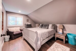 A bedroom with a bed, a nightstand, a dresser, a window with blinds, a bench, and a trash bin. - The Wright's Coach House  (Elora)