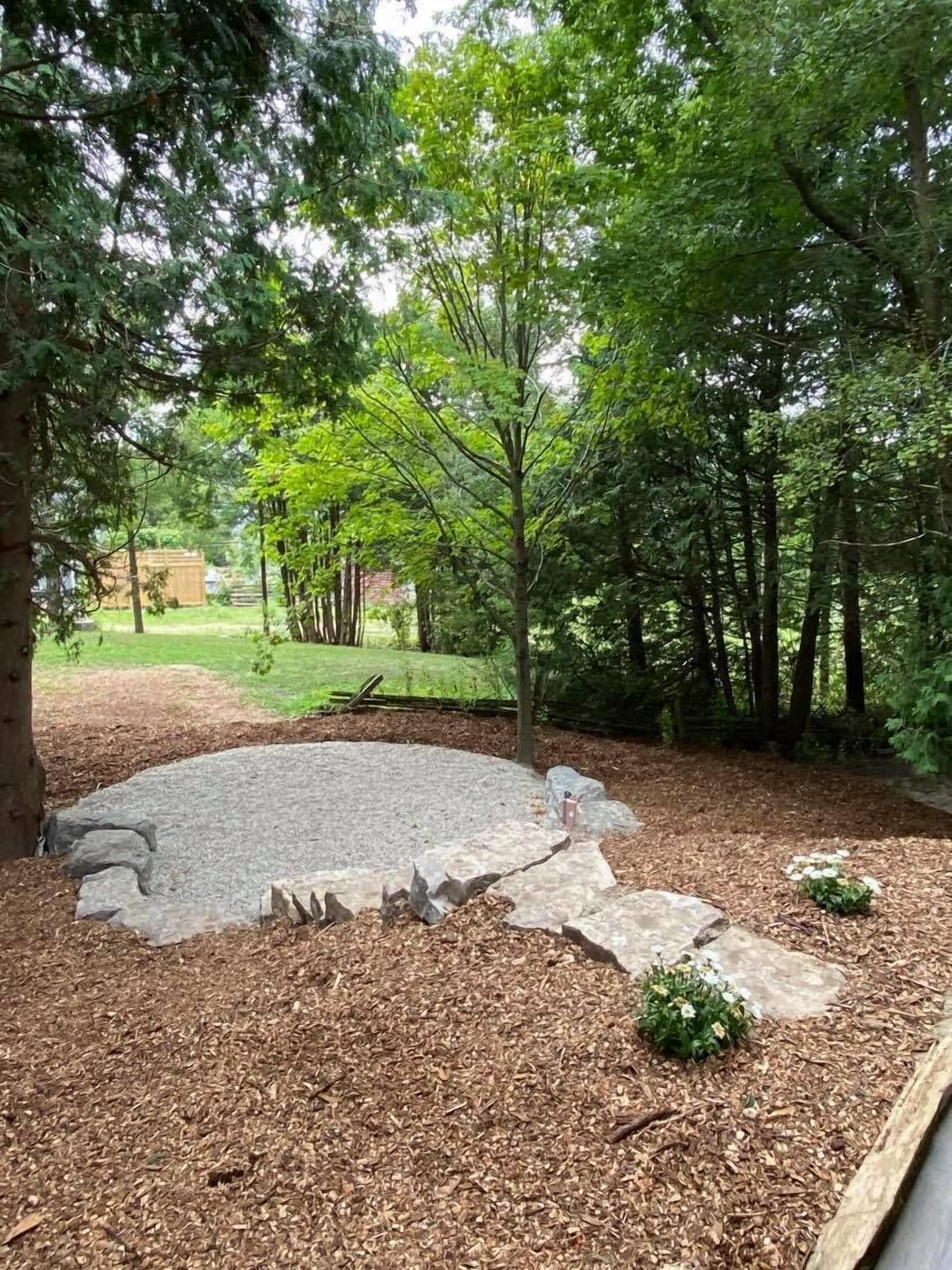 A circular stone patio surrounded by mulch and small plants, with trees in the background.